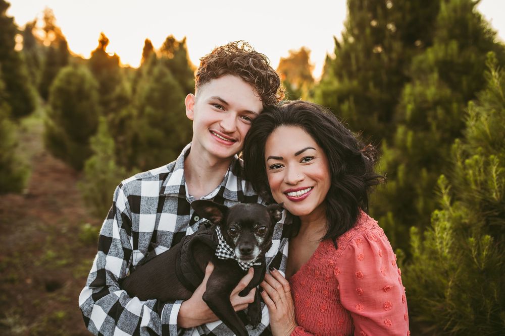Two happy people hold a small black dog while posing at a Christmas tree farm during golden hour sunset.