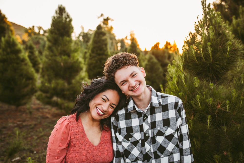 Two people smile together while standing among evergreen trees at sunset in a Christmas tree farm.