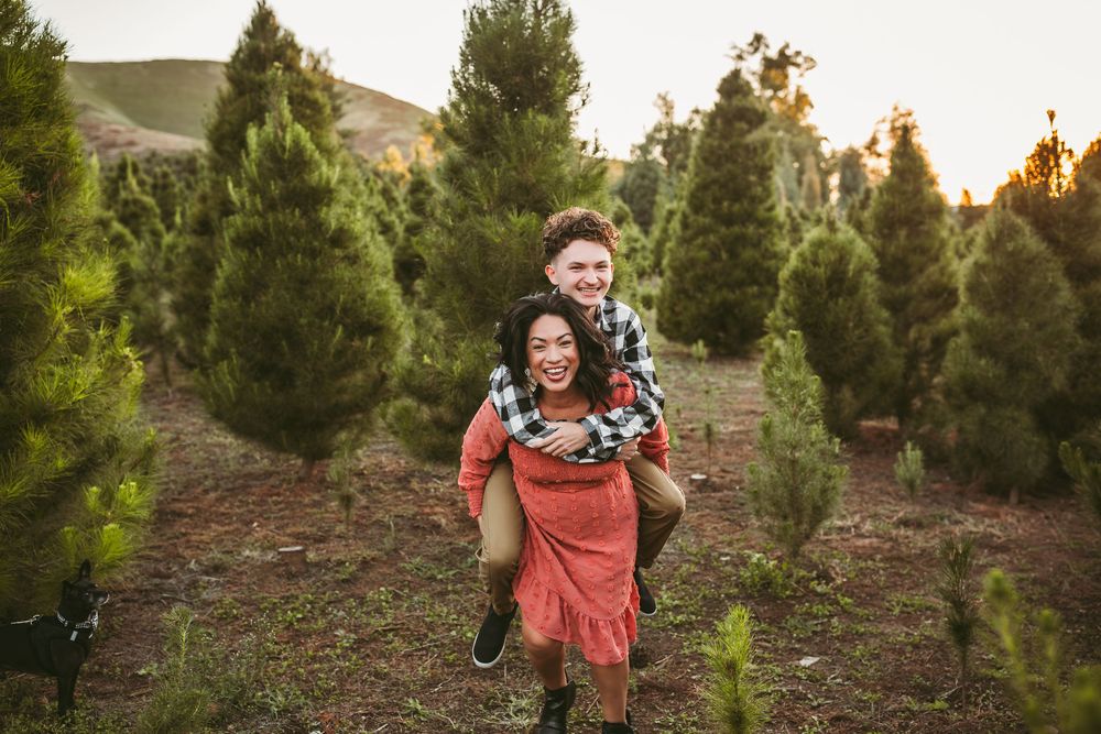 Two people share a playful piggyback moment among evergreen trees at a Christmas tree farm during golden hour sunset.