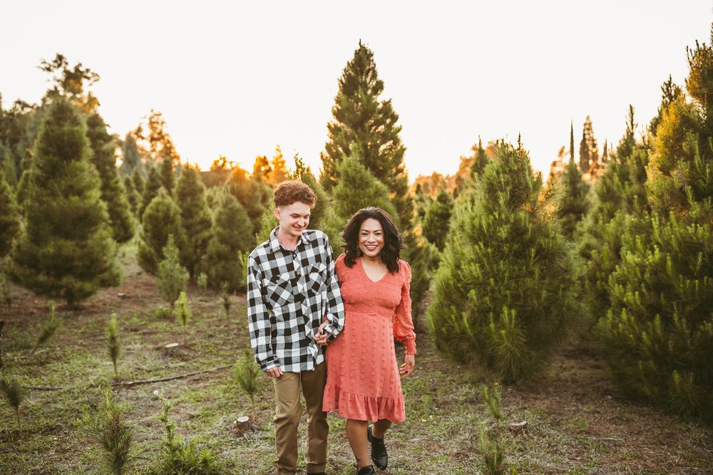A couple stands together at sunset among evergreen trees at a Christmas tree farm wearing casual fall attire.