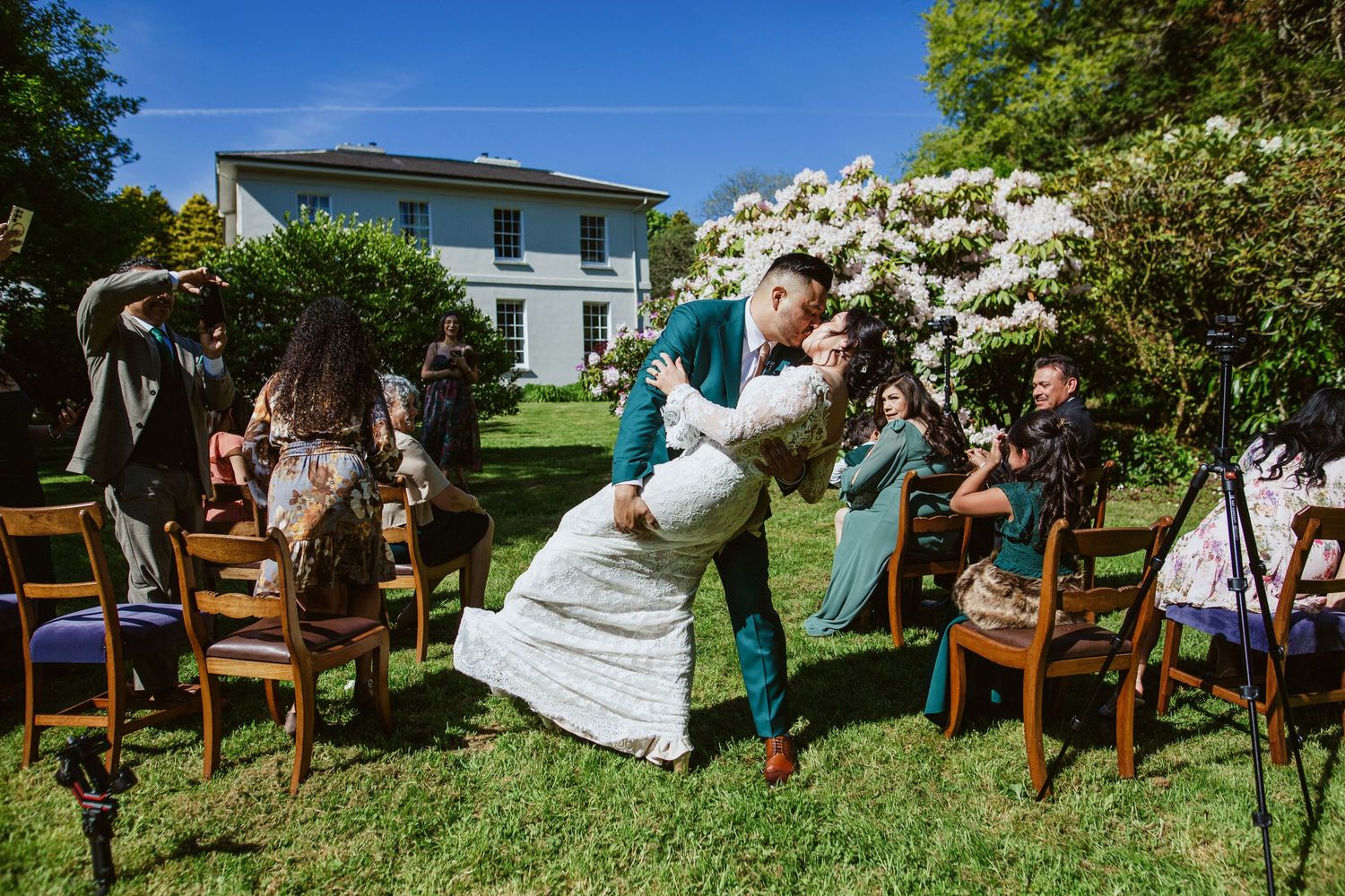Outdoor wedding guests sitting on wooden chairs surrounded by blooming white dogwood trees under a bright blue sky.
