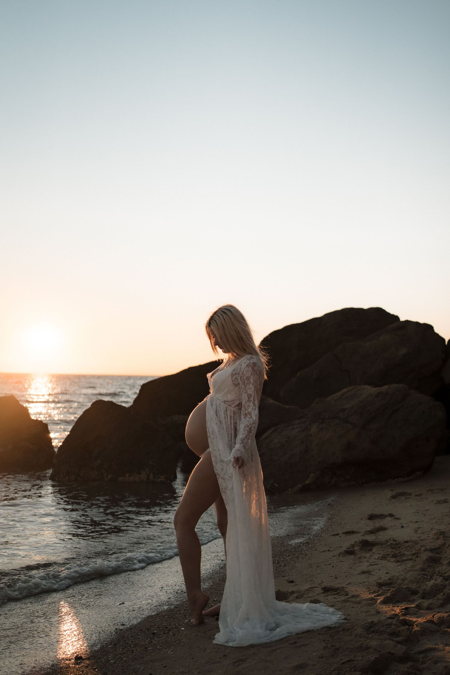 A silhouetted figure in a flowing white lace dress poses on rocky beach during golden sunset over the ocean at Half Moon Bay, Black Rock