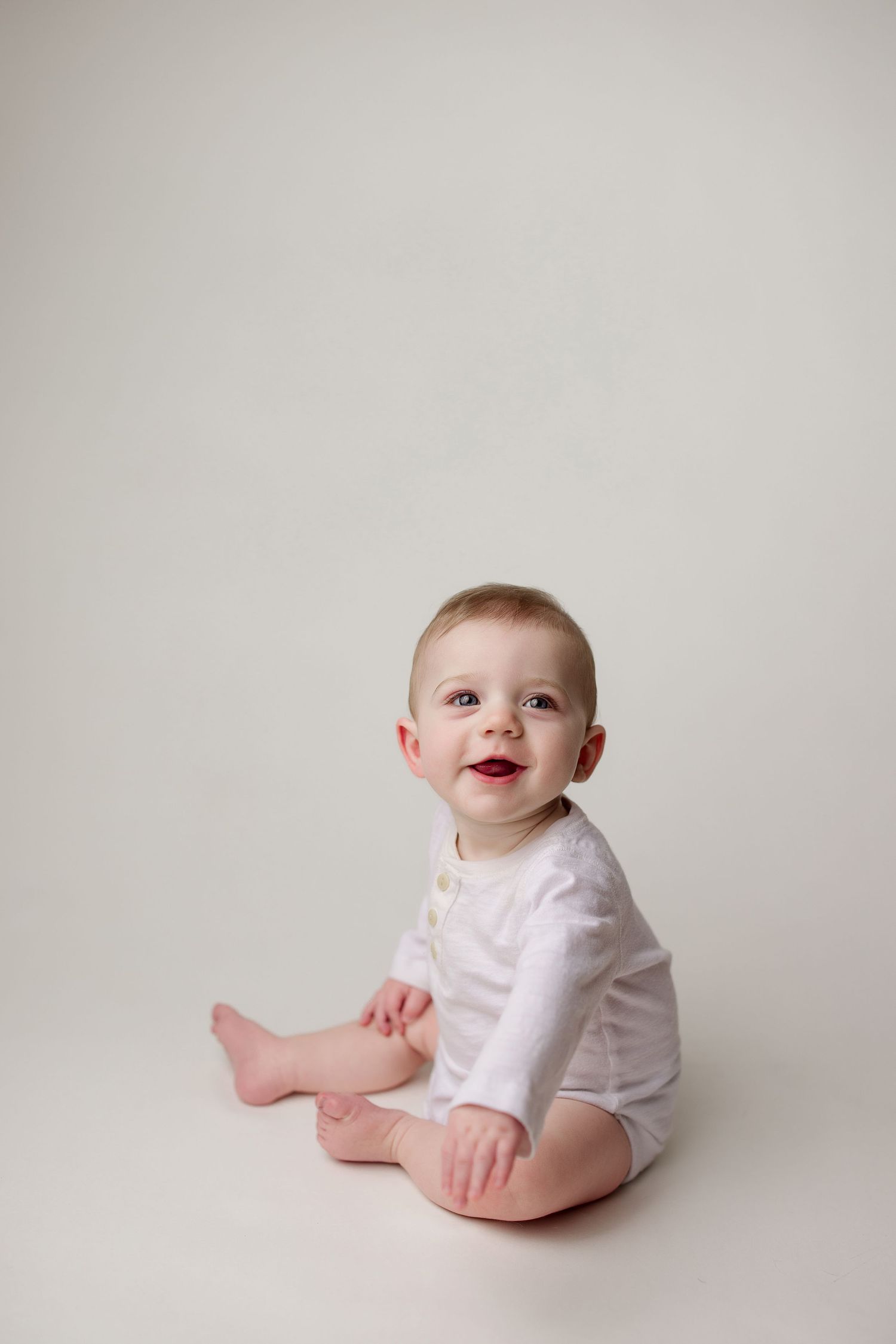 Baby wearing white onesie sits on light seamless background looking up with a happy expression.