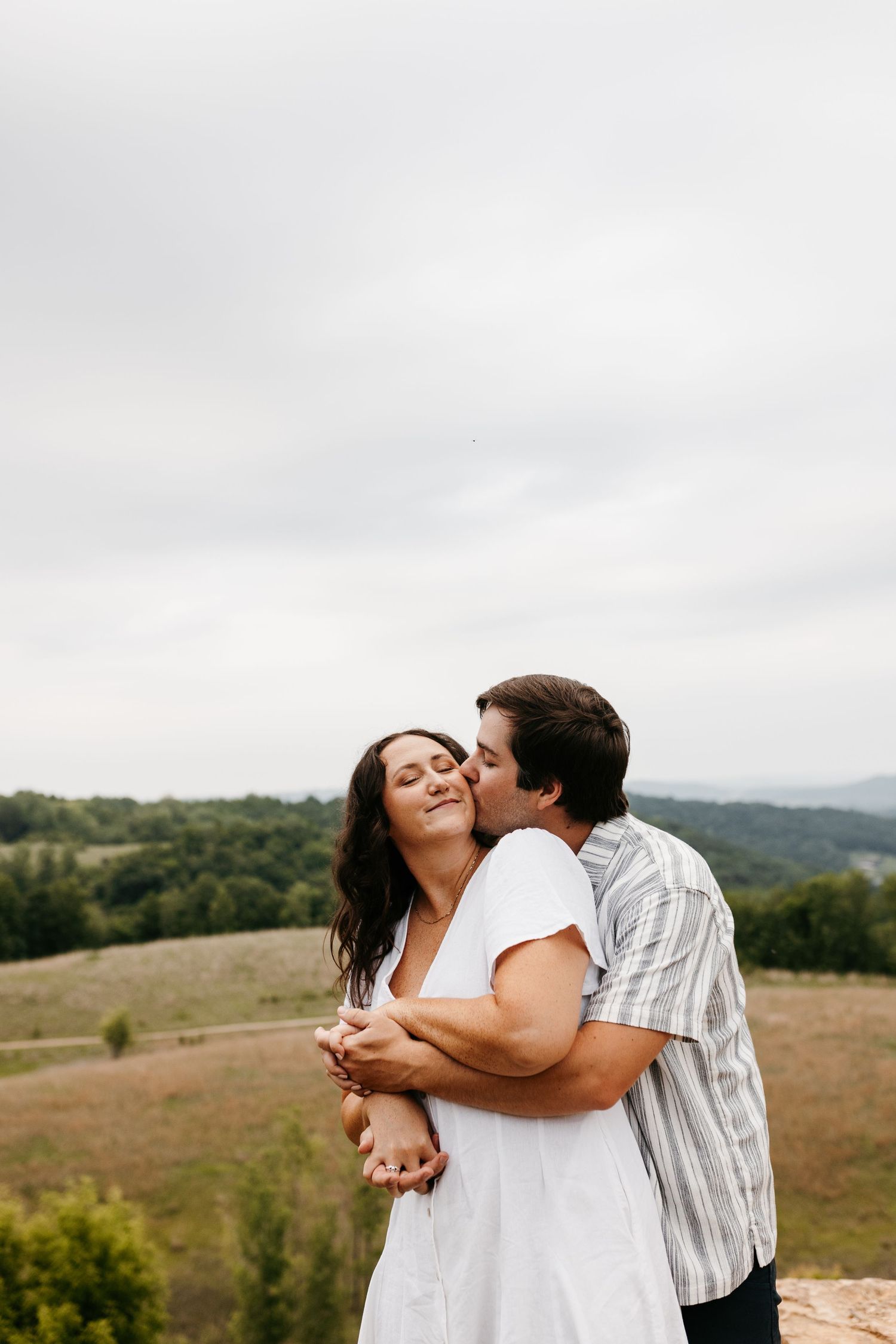 Couple embracing and laughing together on a hilltop overlooking scenic rolling countryside on a cloudy day.