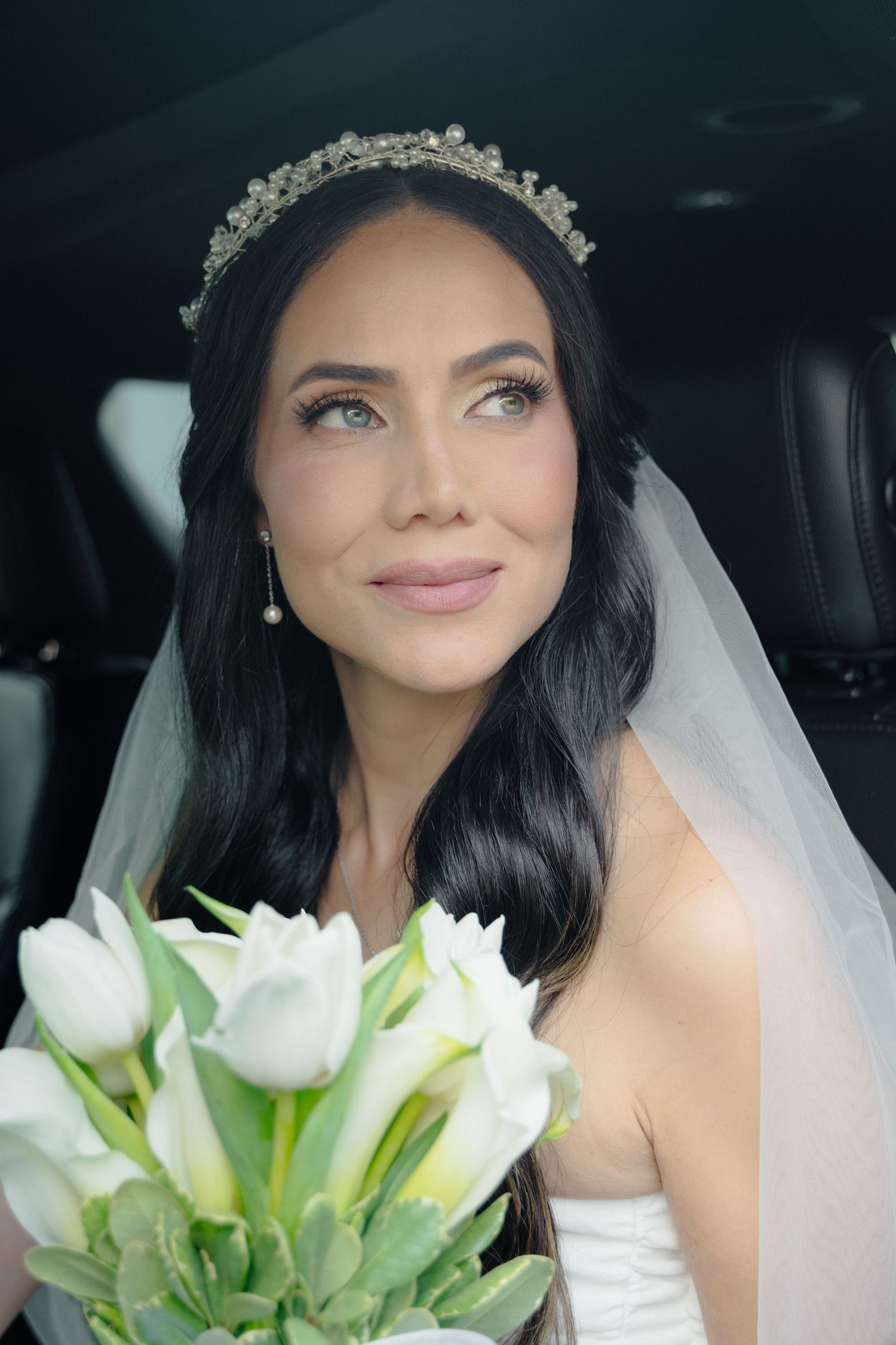 A bride in a white wedding dress holds a bouquet of white tulips while sitting in a car.
