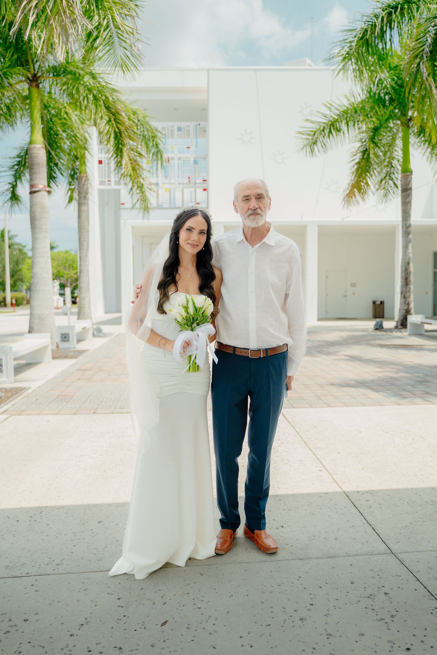 A couple in classic wedding attire poses beneath towering palm trees along a bright white colonnade walkway.