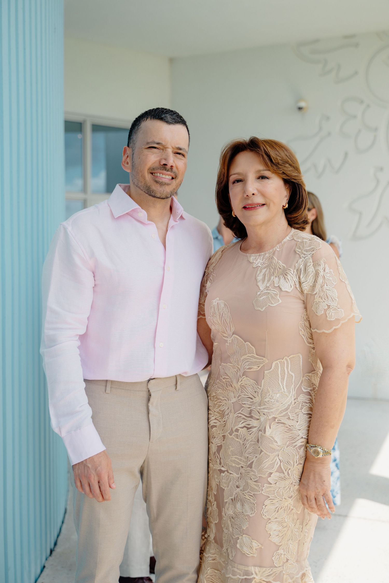 A woman in a beige lace dress and a man in a pink dress shirt and khaki pants stand together against a light wall.