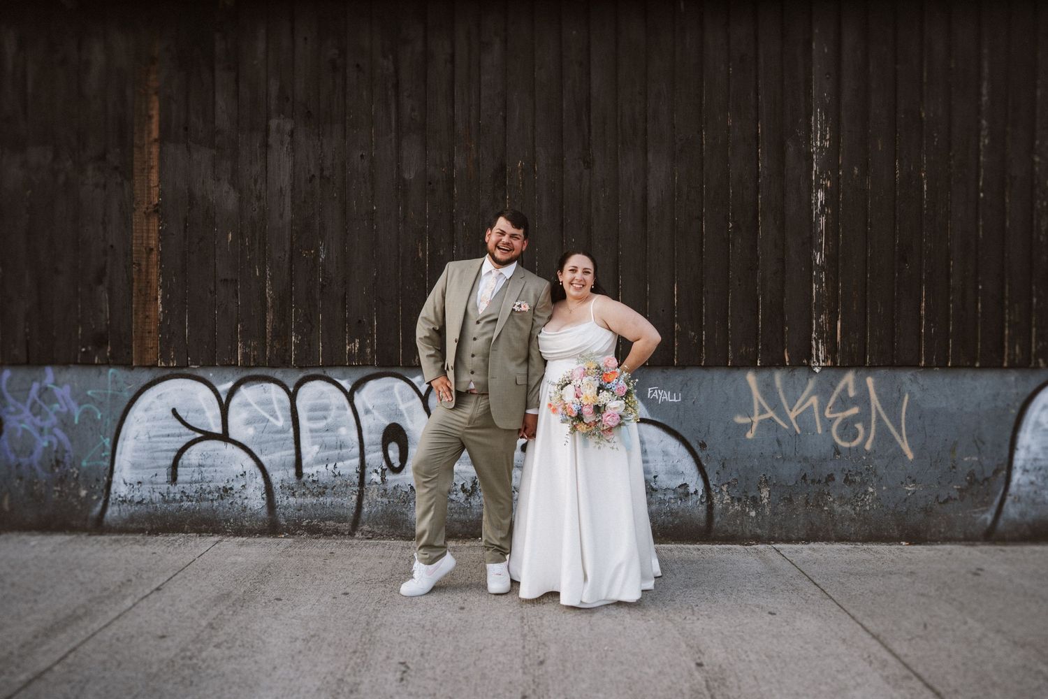 A couple poses together against an industrial wall with graffiti, wearing wedding attire and casual sneakers.
