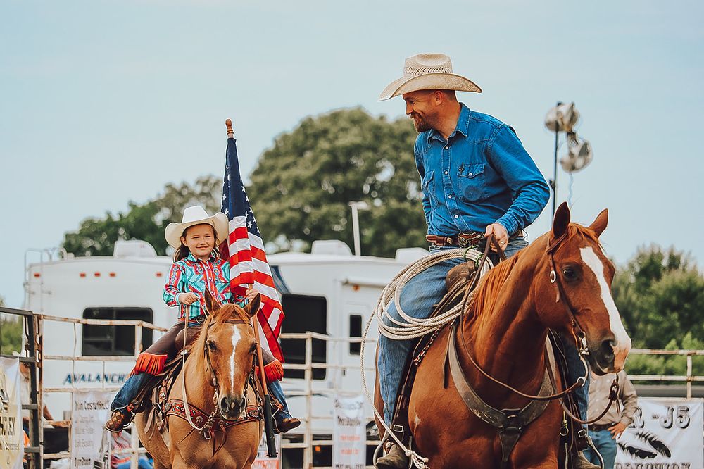 A rider on horseback competing in a western rodeo barrel racing event against a bright outdoor arena backdrop.