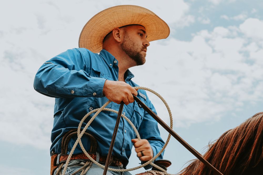 Cowboy in light blue shirt and cowboy hat holds reins while riding horseback against cloudy sky.