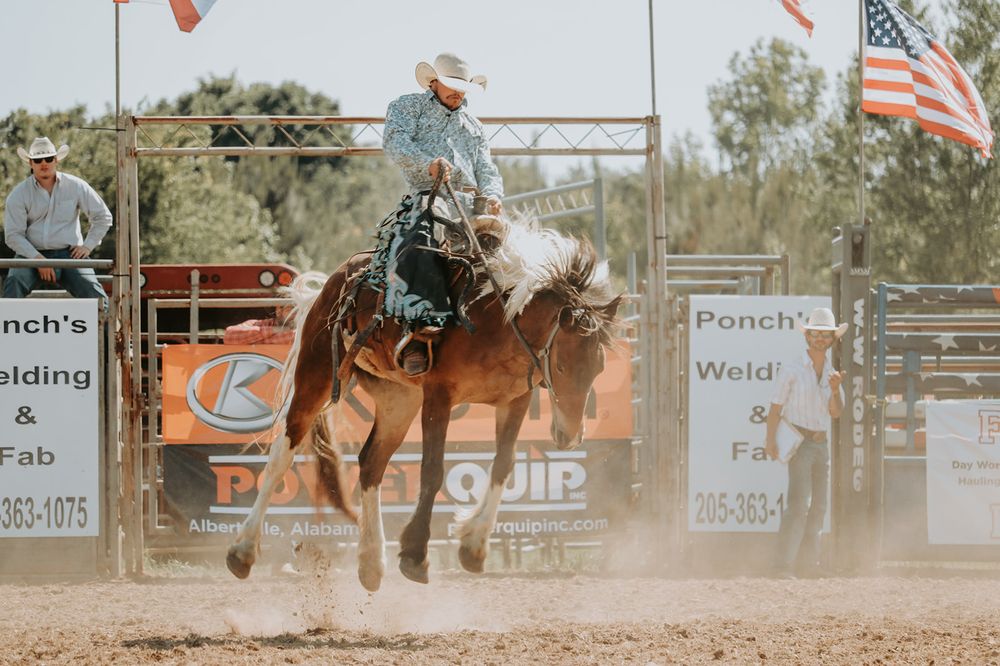 Multiple American flags wave over a dusty rodeo arena during a western competition event.