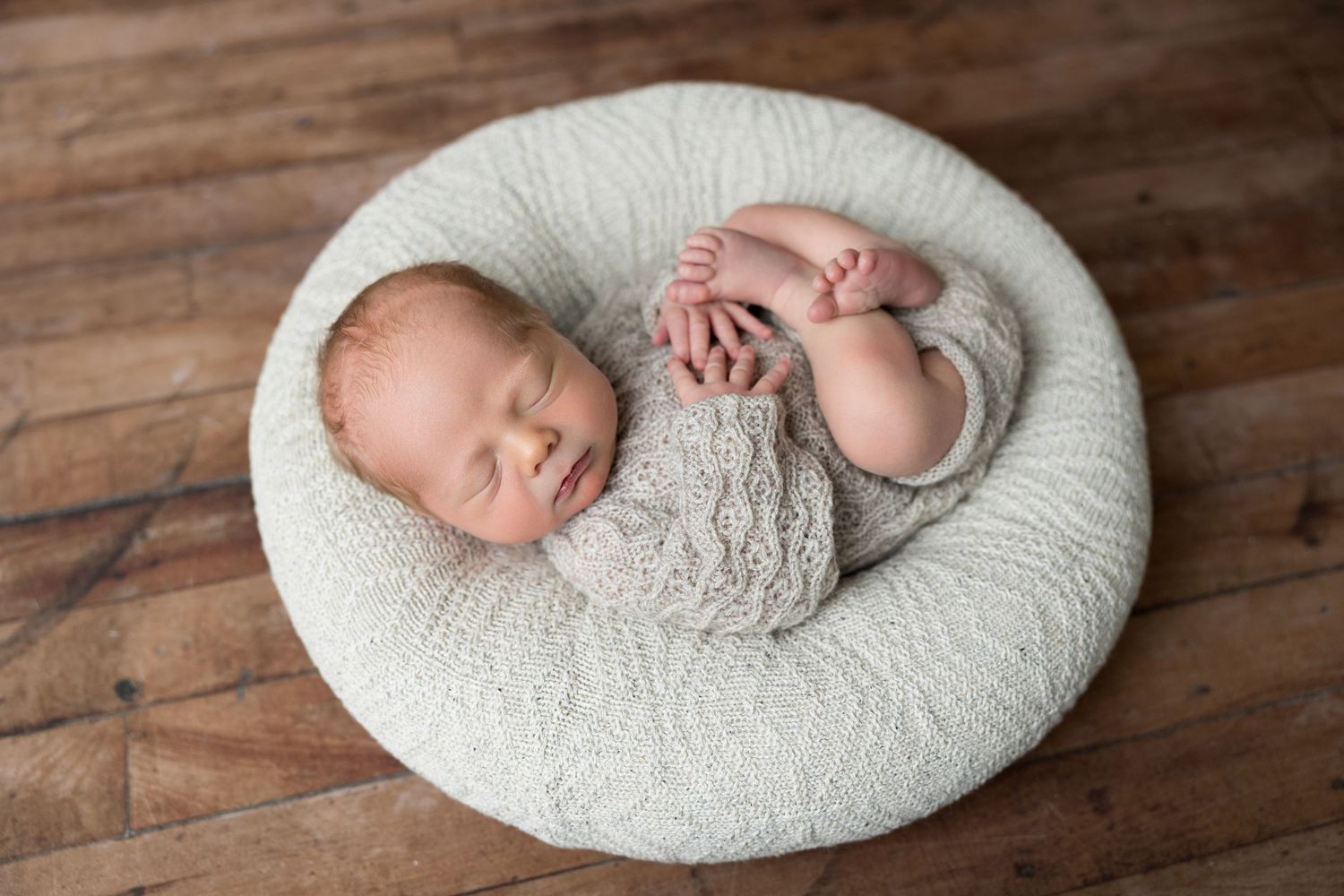 Newborn baby sleeping peacefully in a round white cushion on wooden floor during professional photo session.