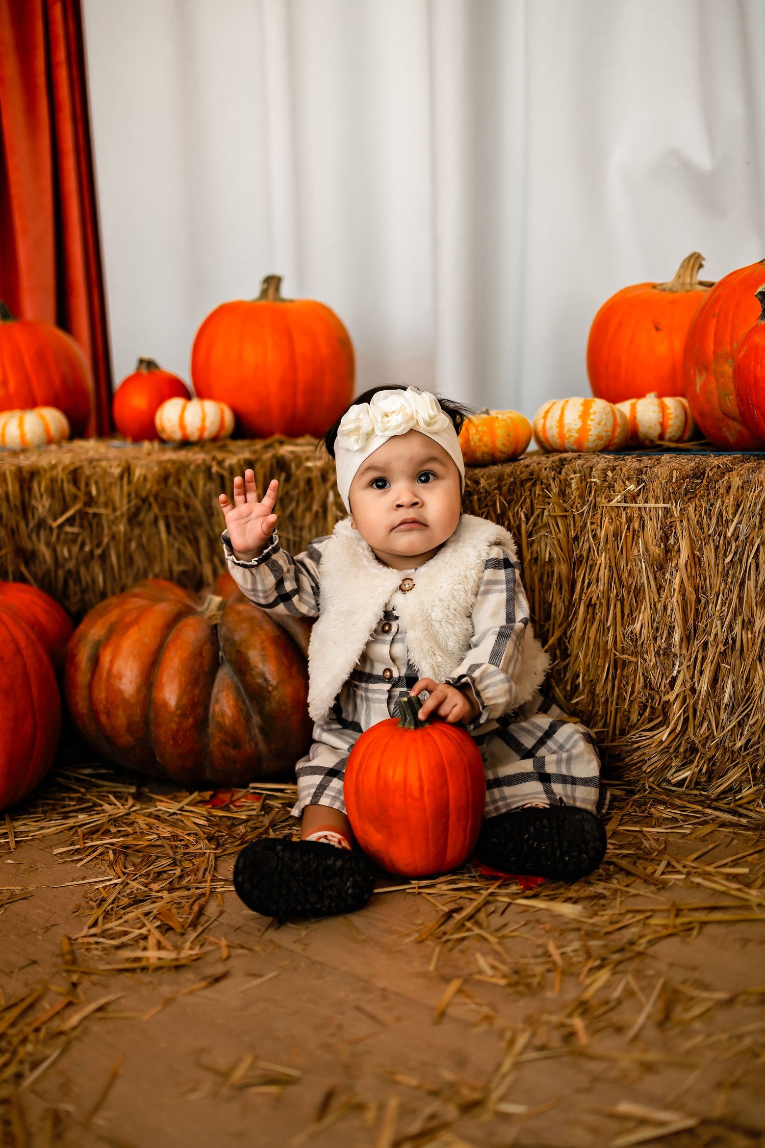 Small baby in white outfit sitting on hay bales with autumn pumpkins in festive fall photo setting.