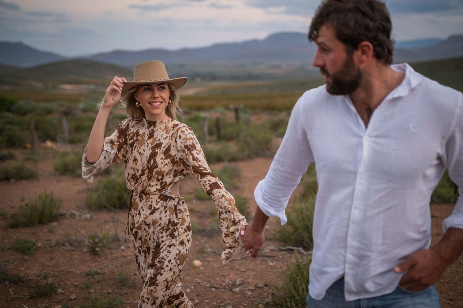 A couple enjoys a playful moment in the desert landscape at sunset, with mountains visible in the background.