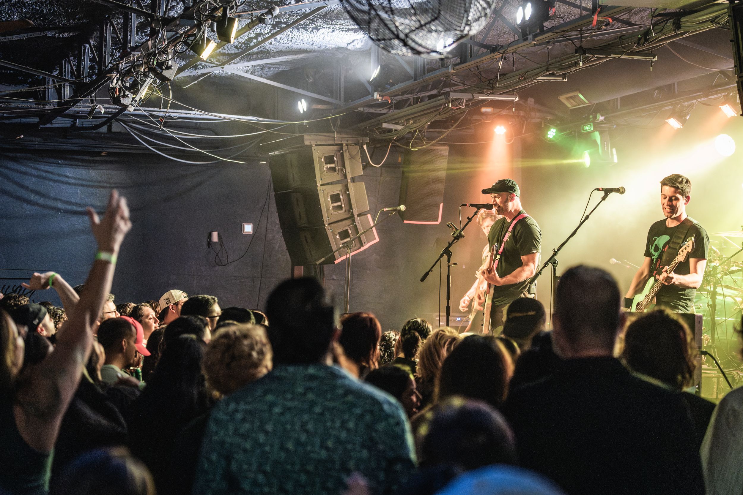 Musicians perform on stage at an intimate concert venue while an energetic crowd raises hands in the dark room.