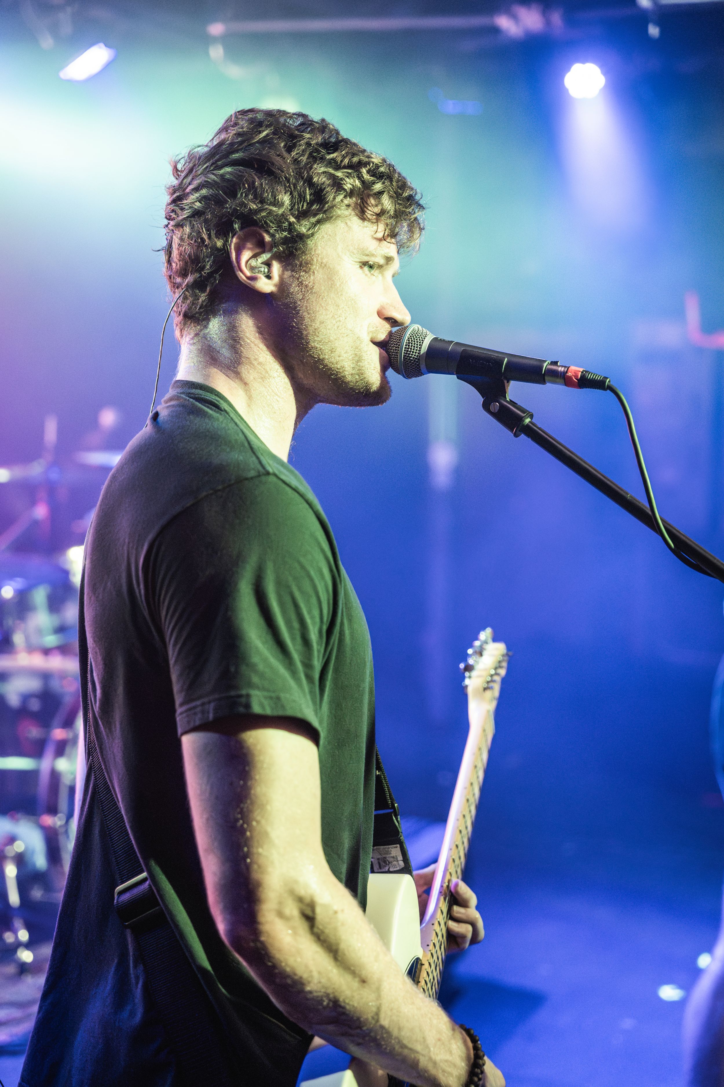 A performer in a black t-shirt plays guitar on stage with dramatic blue lighting and fog effects during a live concert.