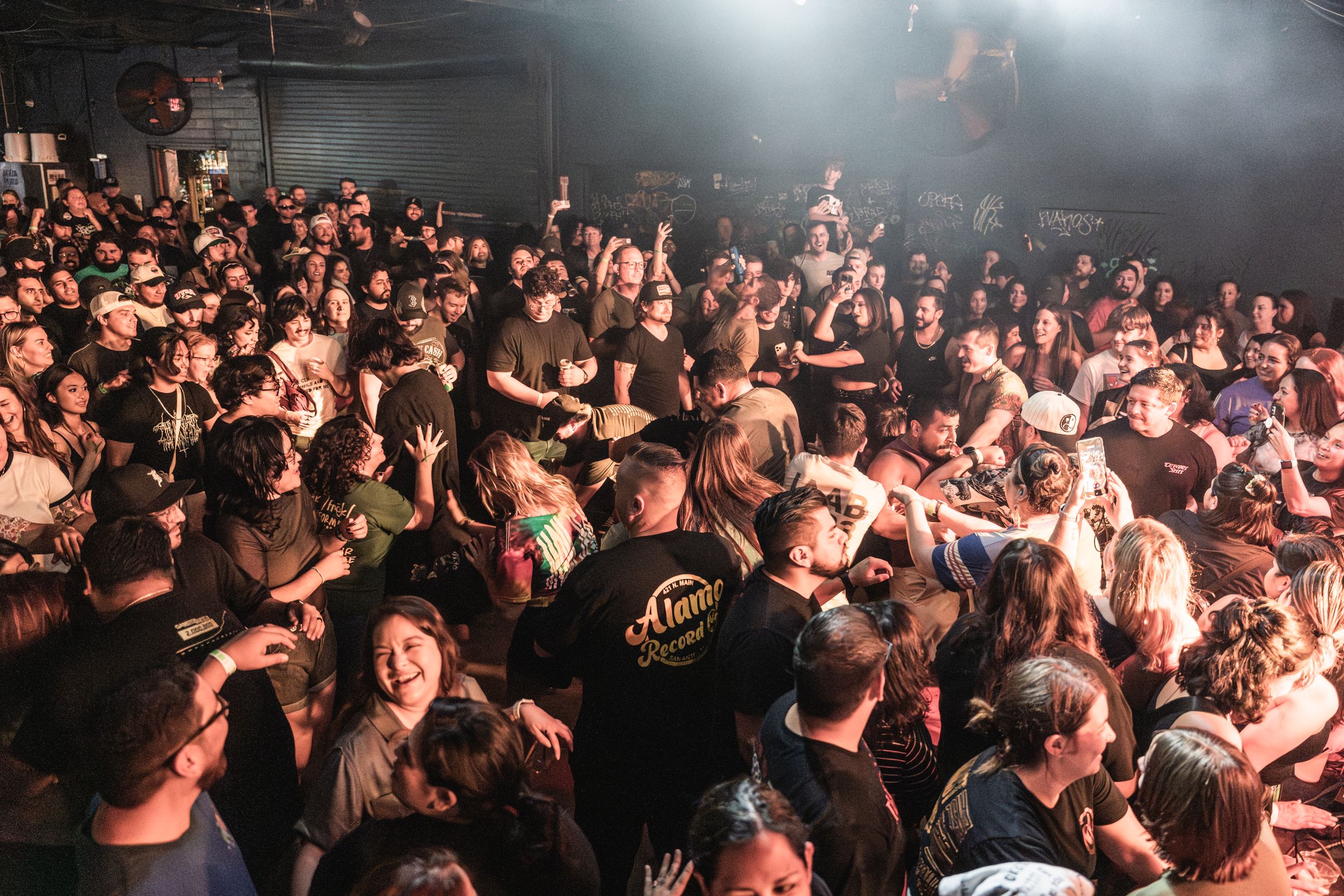 An energetic crowd of concert-goers fills a dark venue with raised hands and dramatic lighting from above.
