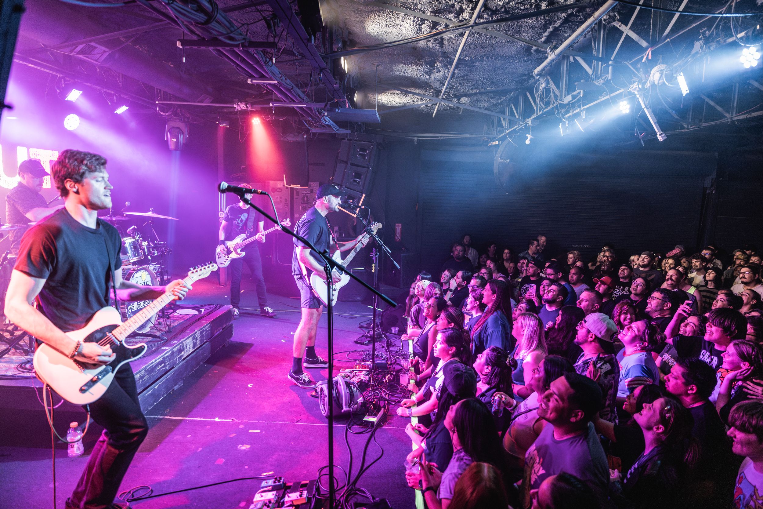 Concert crowd watches energetic performance in purple-lit venue as performers play instruments on stage.