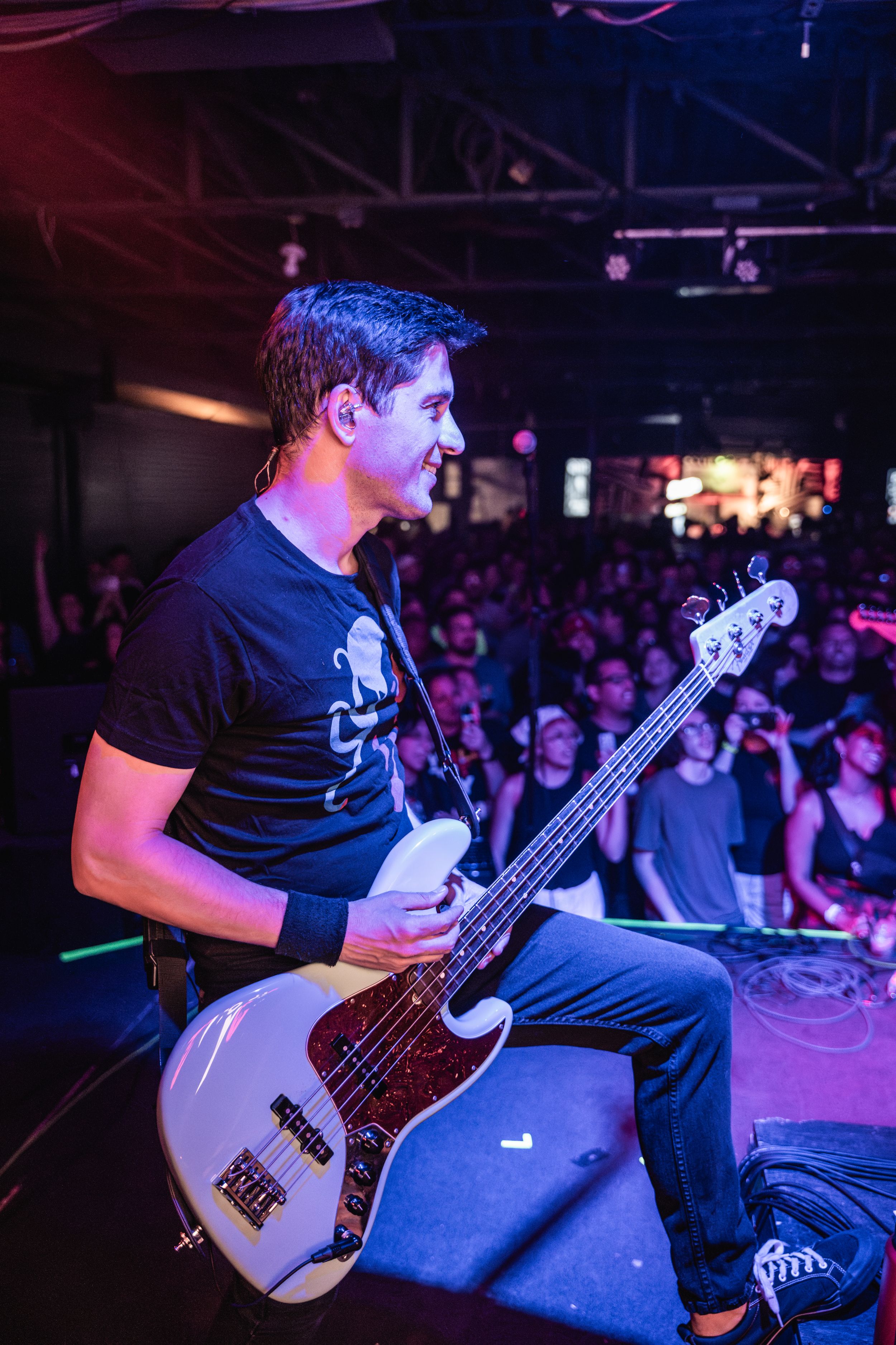 Bass guitarist in a black t-shirt performs on stage at a concert venue with blue and red lighting.