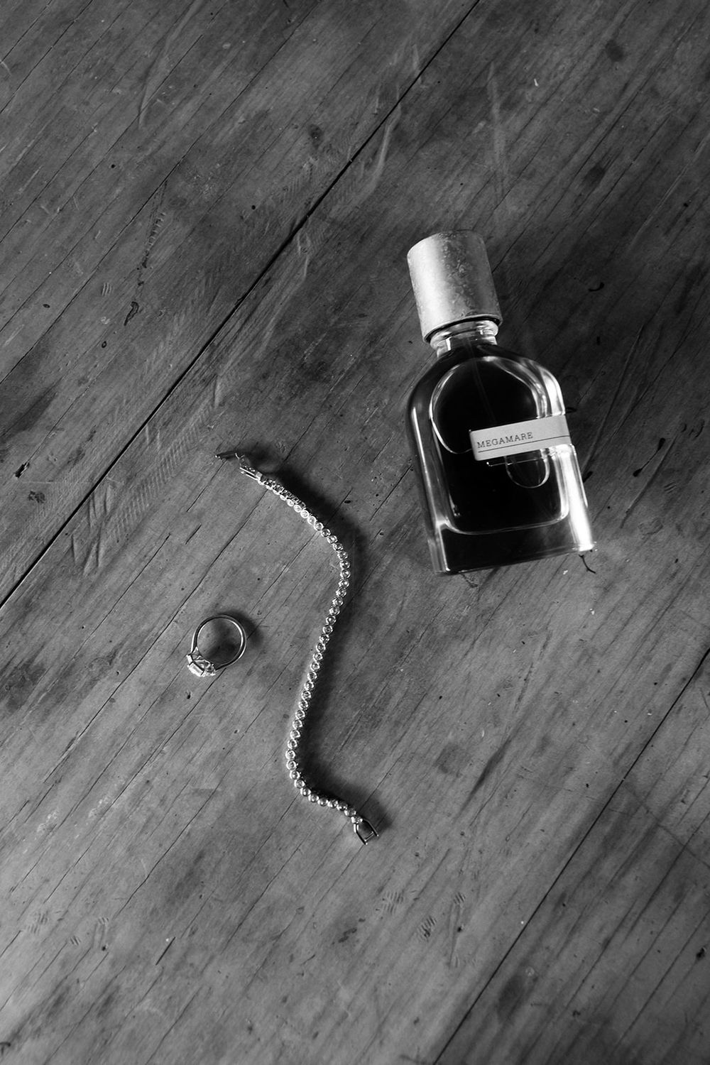 Black and white photo of a perfume bottle and bracelet on a wooden floor, shot from above.