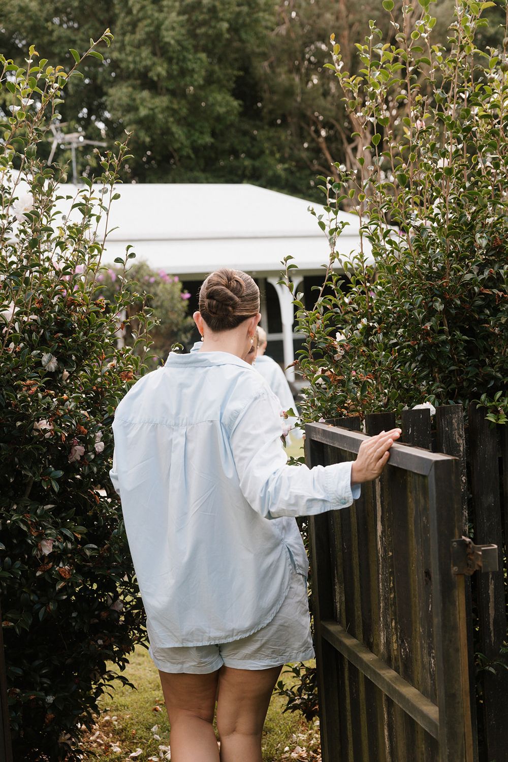 A figure in a light-colored shirt and shorts walks along a wooden fence in a garden setting.