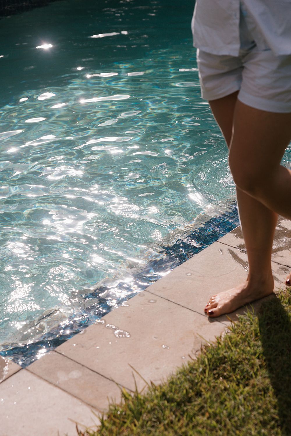 Bare feet stand at the edge of a shimmering swimming pool on a sunny day.
