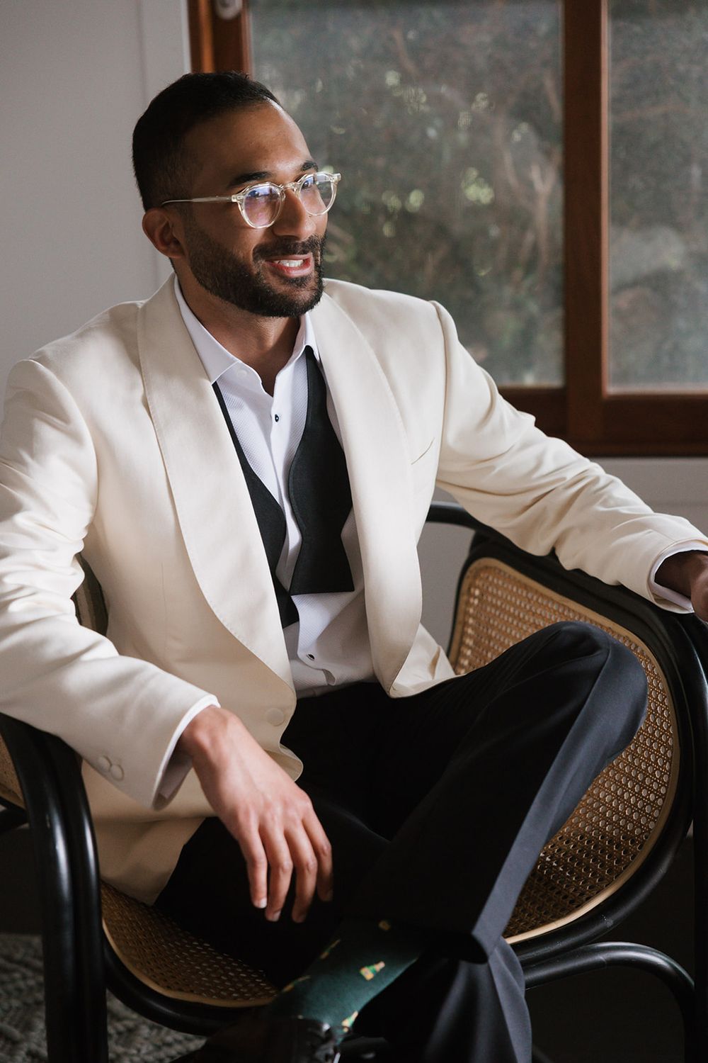 A stylish person wearing glasses and a white blazer sits relaxed in a cane-backed chair by a window.