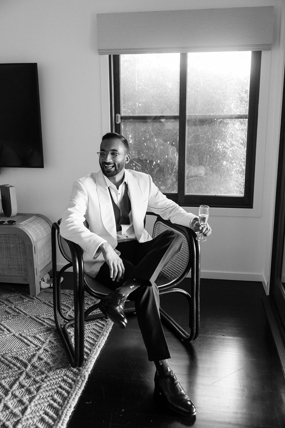 Black and white photo of someone in a white blazer and dark pants sitting in a modern chair in a minimalist room.