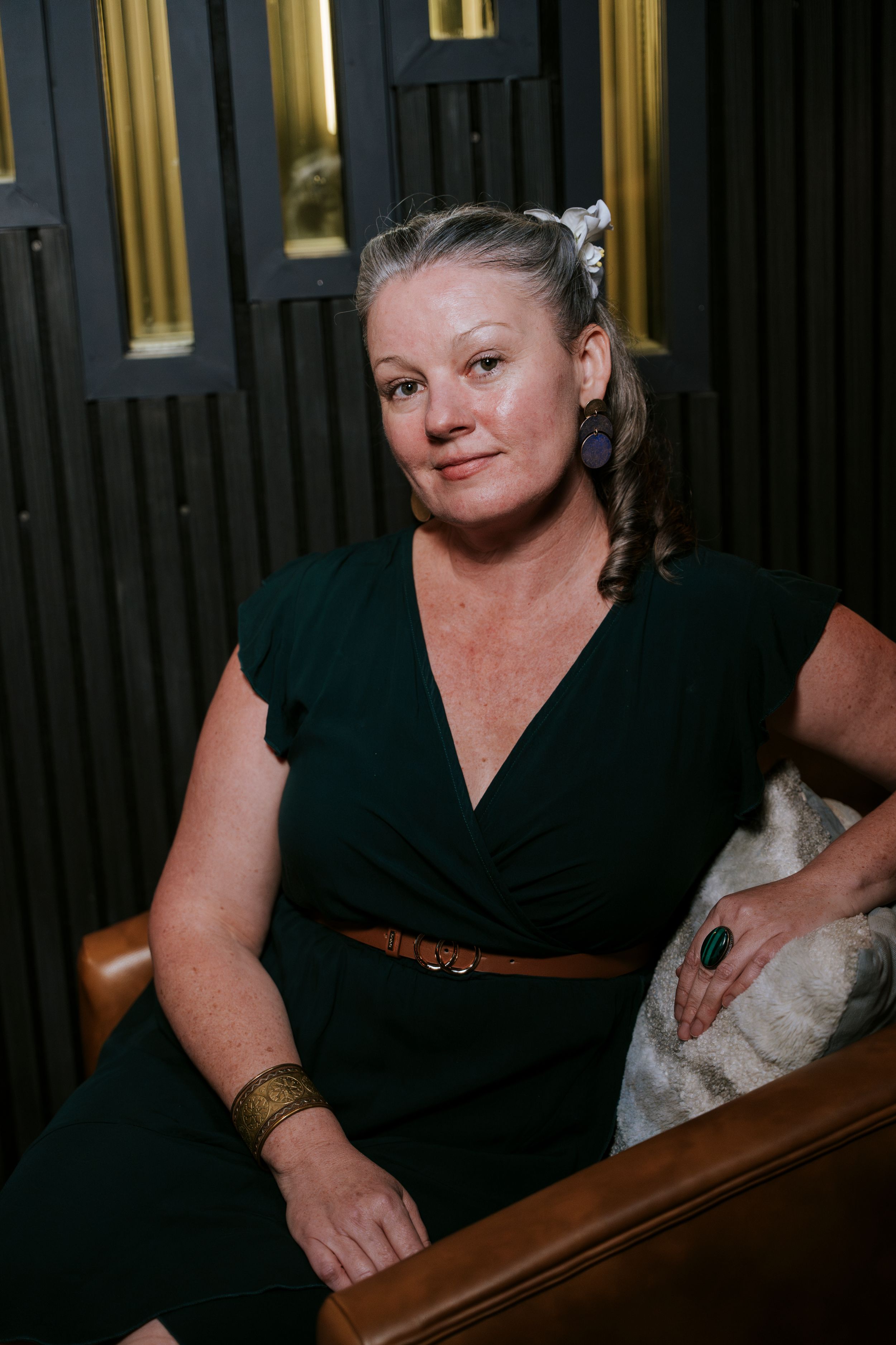 A person wearing a black dress with a brown belt sitting at a wooden table in dim lighting.