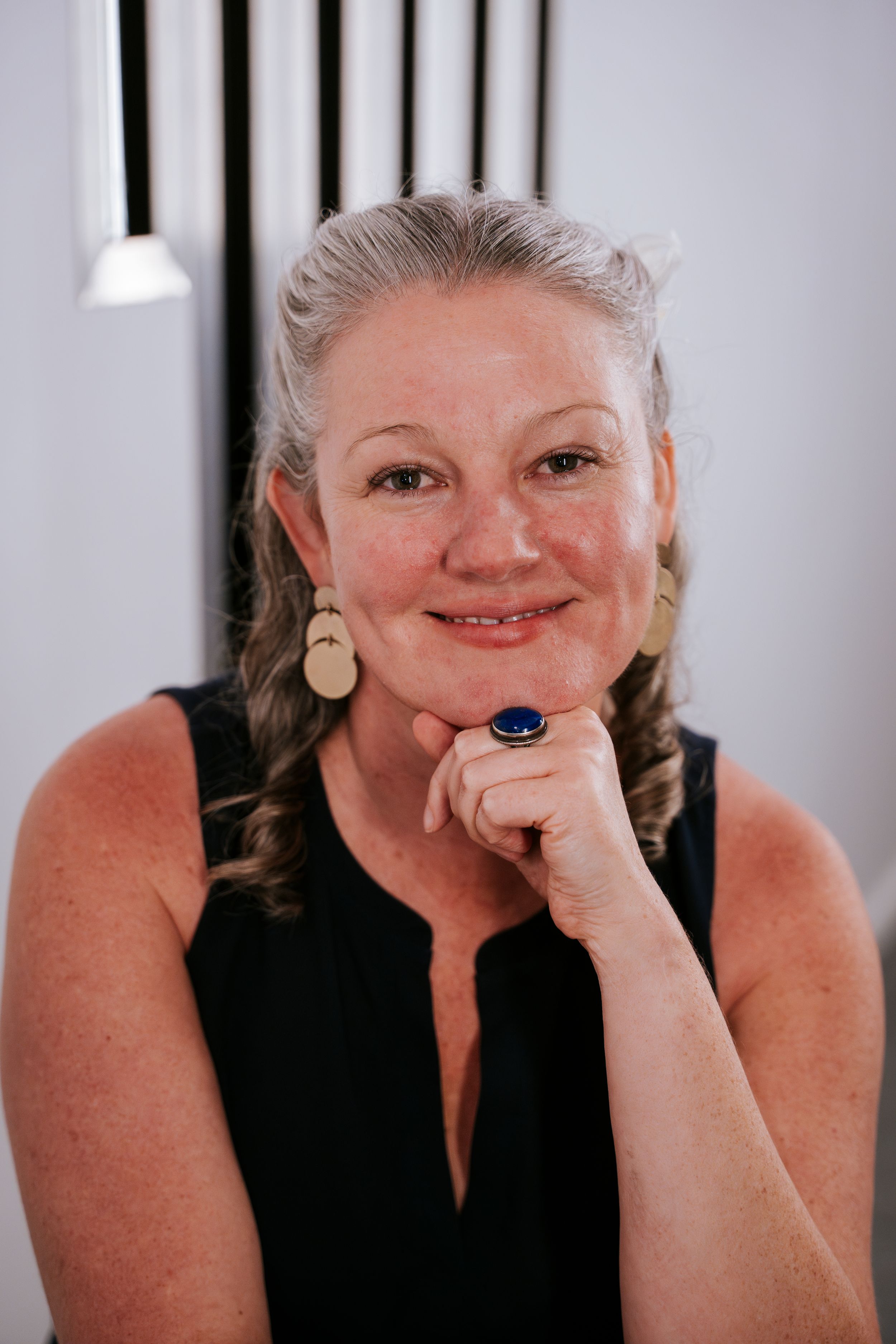 A person wearing a black sleeveless top smiles warmly while wearing a statement blue ring and gold drop earrings.