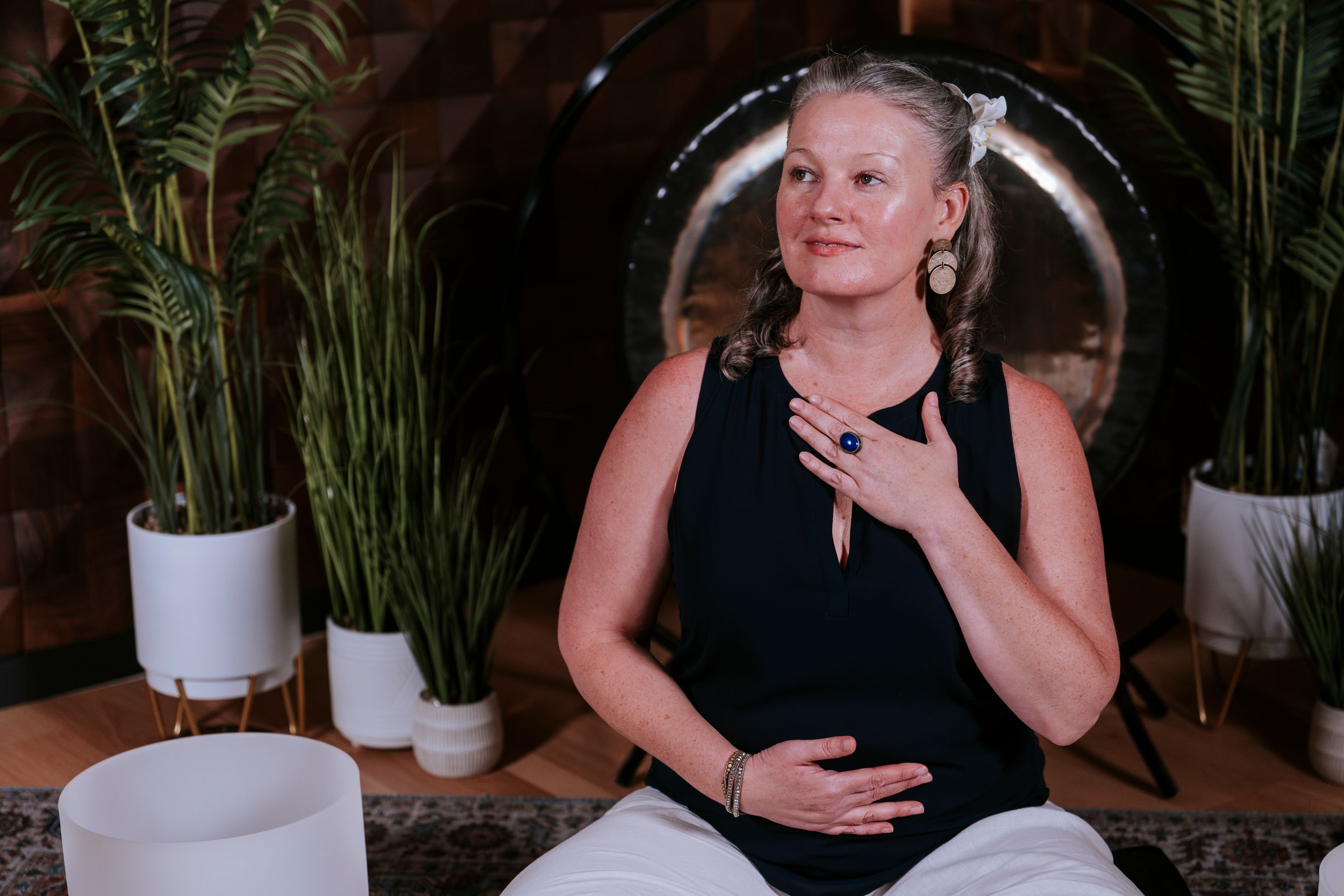 Person in black tank top sits in meditation pose with indoor plants and white ceramic pots in background.