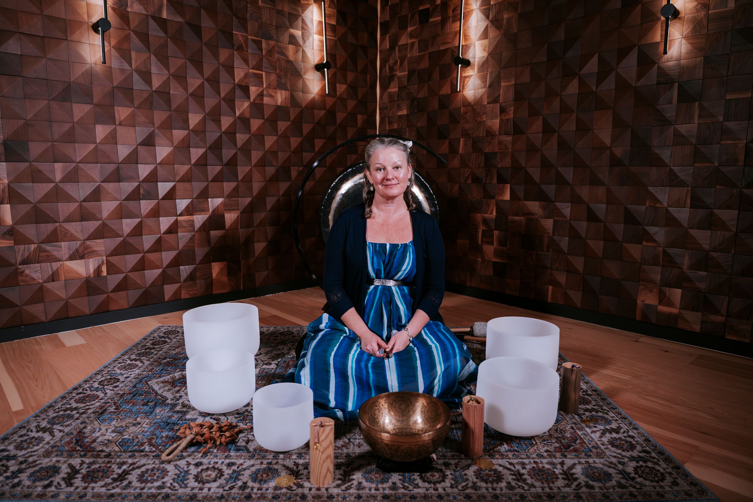 A person in a blue dress sits on the floor surrounded by white crystal singing bowls and a geometric copper wall.