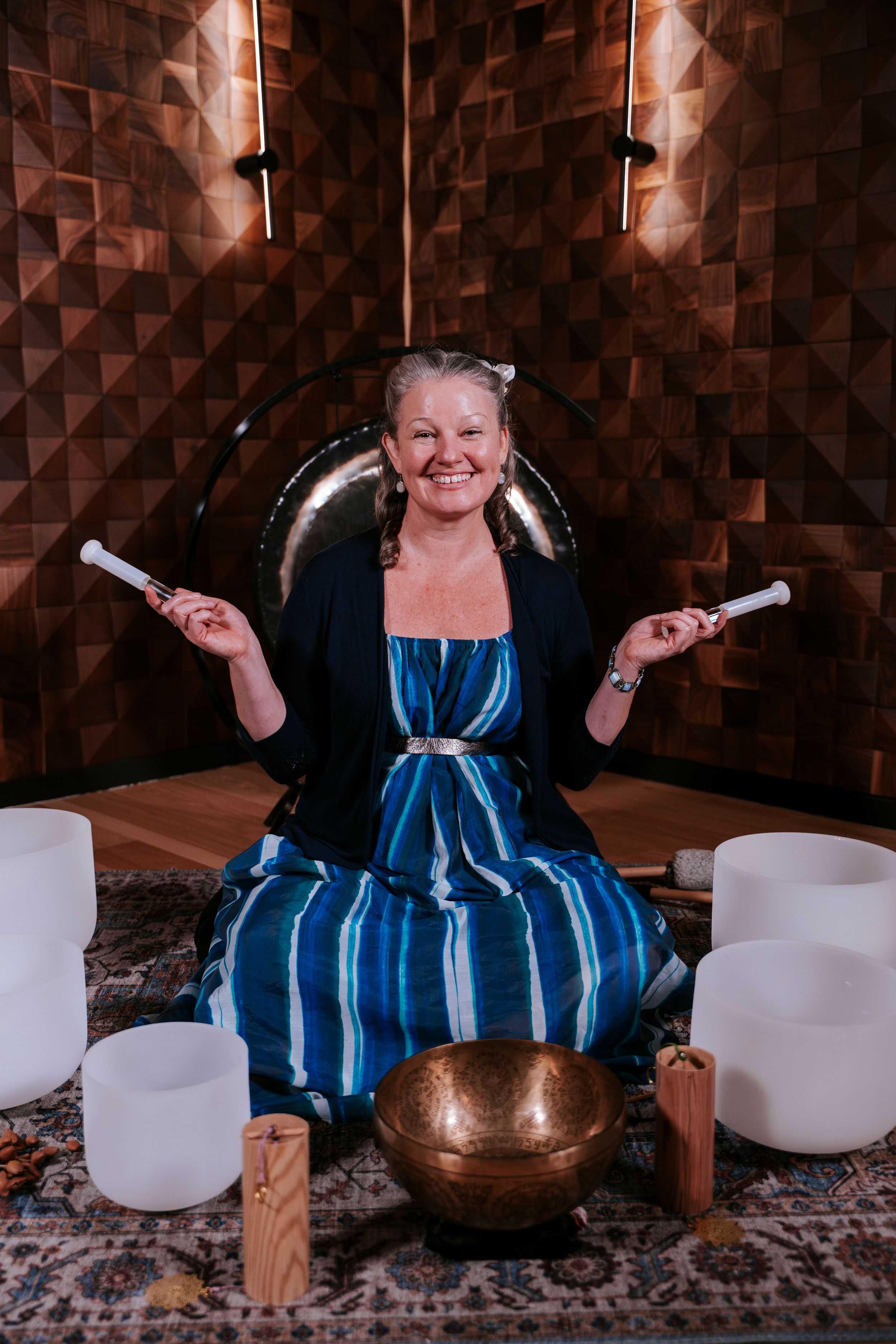 Person in blue striped dress sits among crystal singing bowls and healing instruments during sound meditation session.