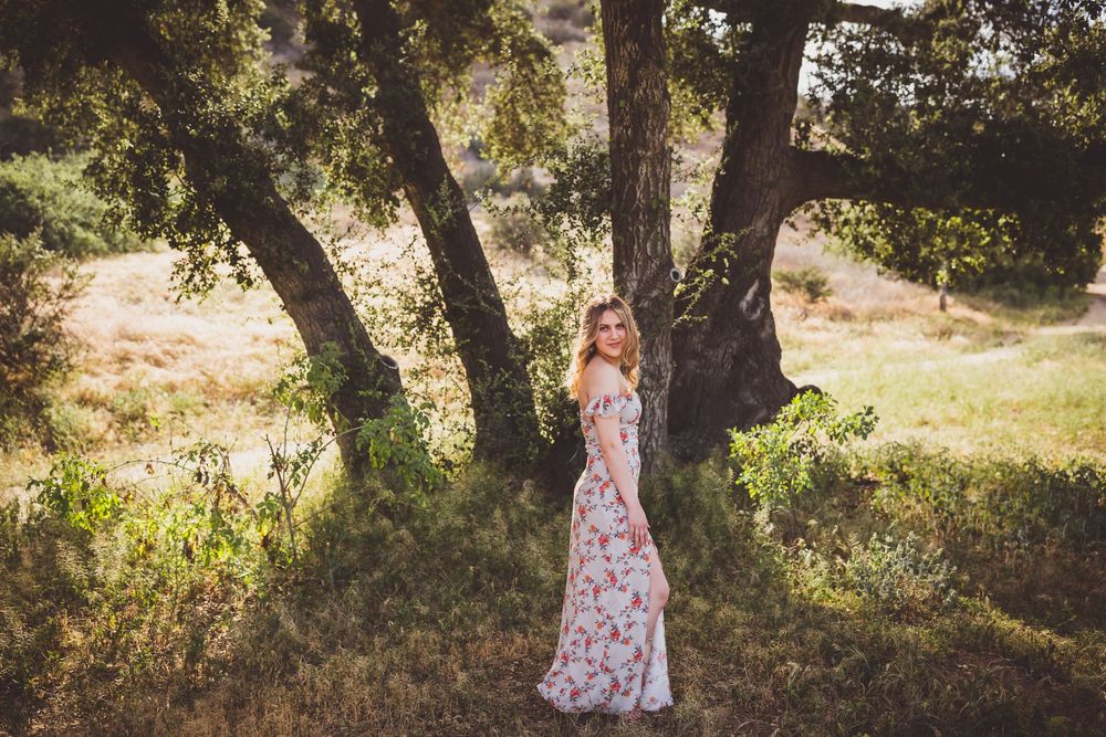 A dreamy woodland photo session features someone in a floral dress walking among sunlit oak trees in a natural setting.