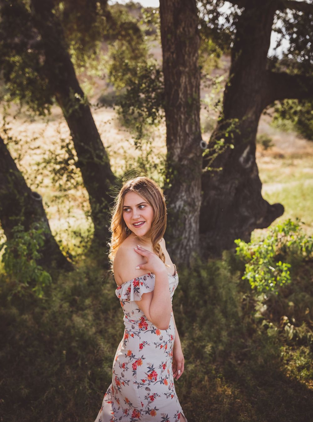 A person in a floral sundress poses gracefully in a sunlit wooded area during a summer photoshoot.