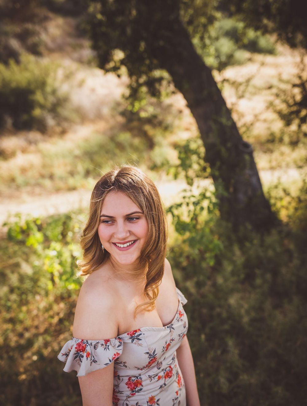 A woman in a floral off-shoulder dress poses outdoors in natural sunlit surroundings.