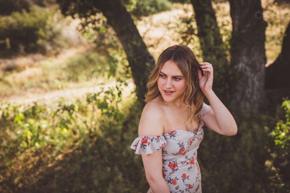 Woman in floral dress sitting gracefully outdoors in golden sunlight with trees in background.