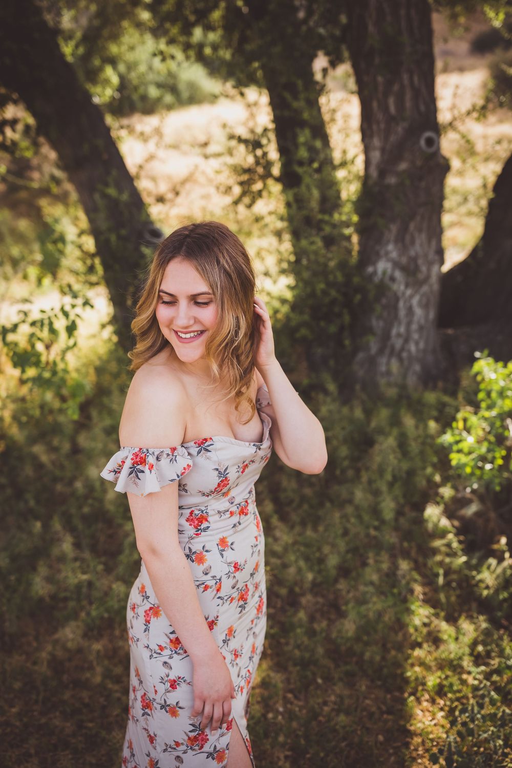 A woman in a floral off-shoulder dress smiles joyfully while standing in a sunlit wooded area.
