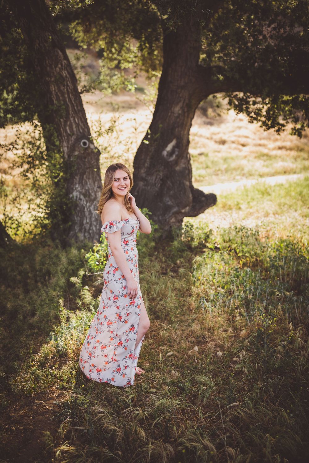 A floral dress catches sunlight in a dreamy outdoor photoshoot among oak trees and tall grass.