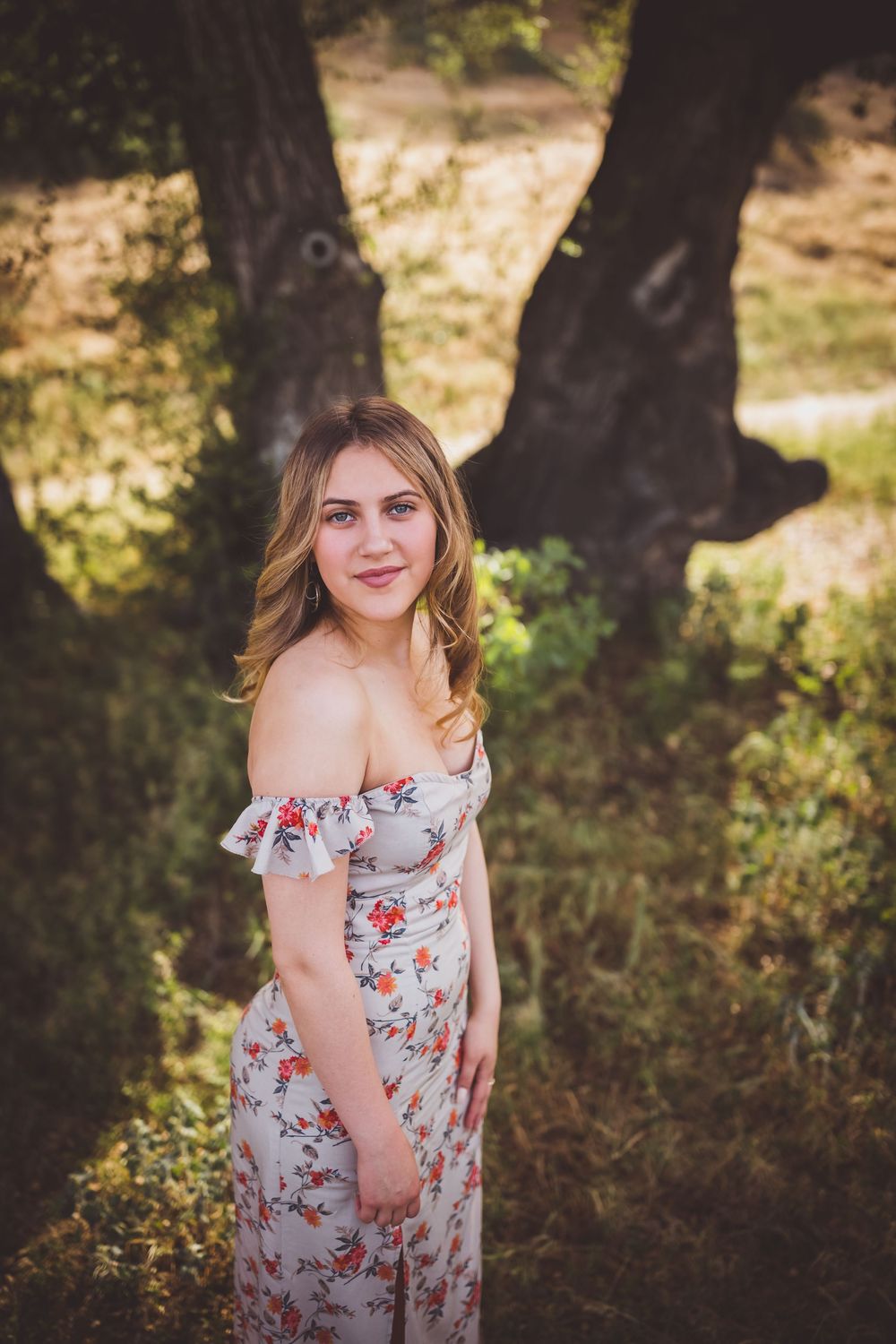 A young girl in a floral off-shoulder dress poses in front of trees with soft natural lighting creating a dreamy outdoor portrait.