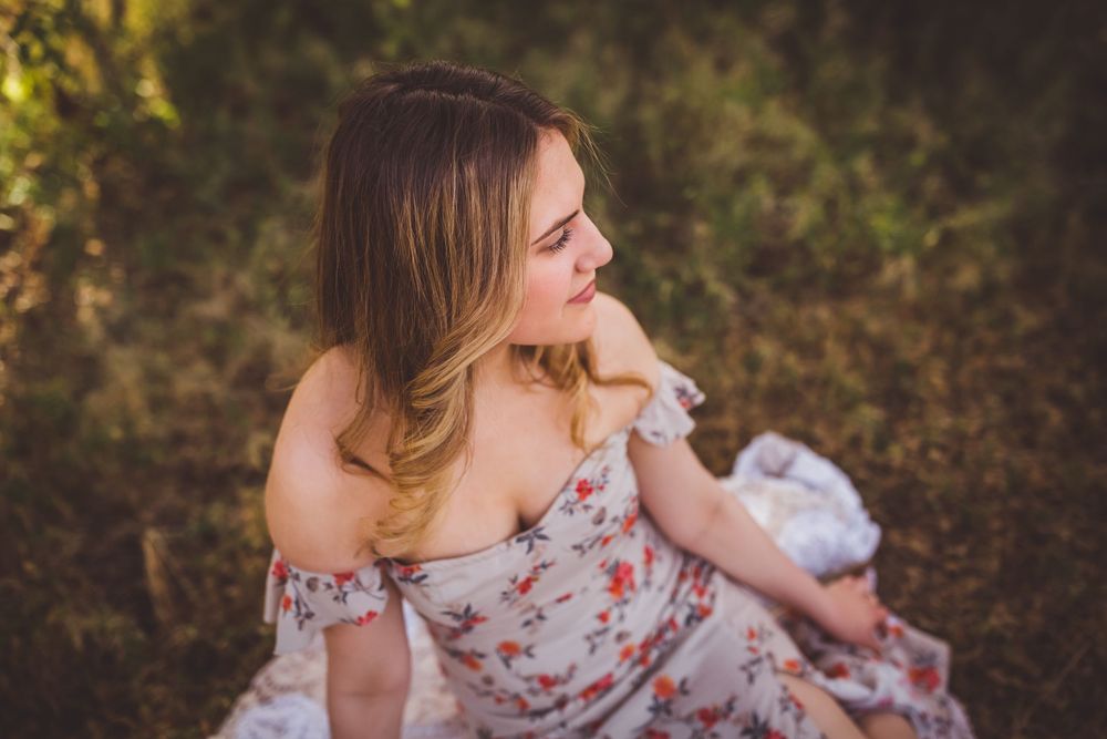 Woman in a floral dress sits on grass in natural outdoor setting with soft golden lighting at sunset.