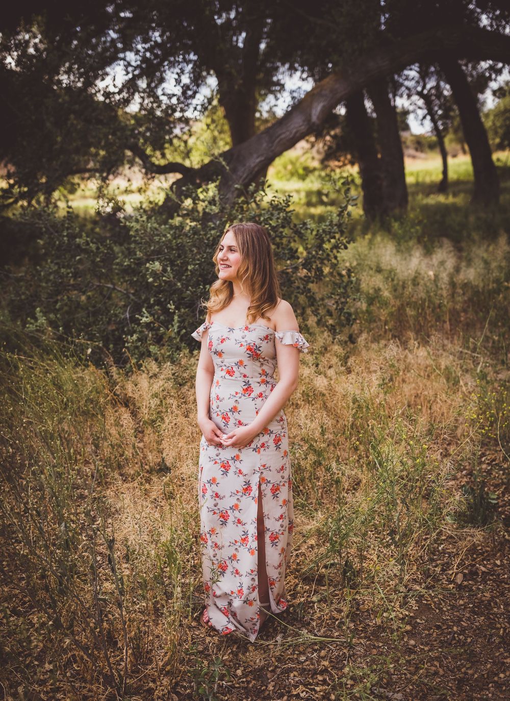 Natural light filters through trees as someone poses in a floral dress against a rustic outdoor backdrop.