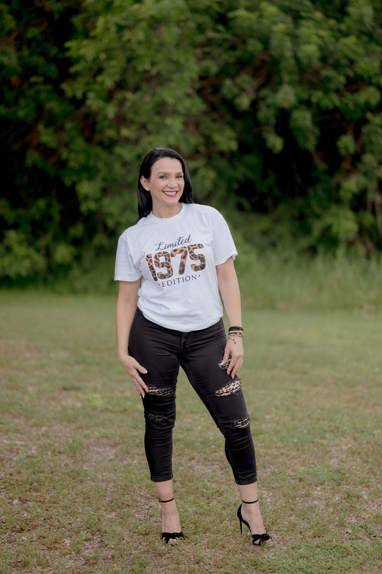 Woman in a white 1975 t-shirt and distressed black jeans poses outdoors in a grassy field with trees in background.