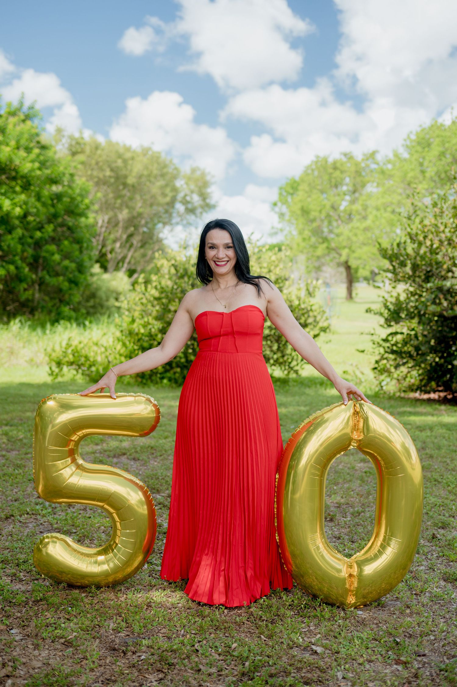 Someone stands outdoors in a red pleated dress next to gold number balloons showing 50 against a green natural background.