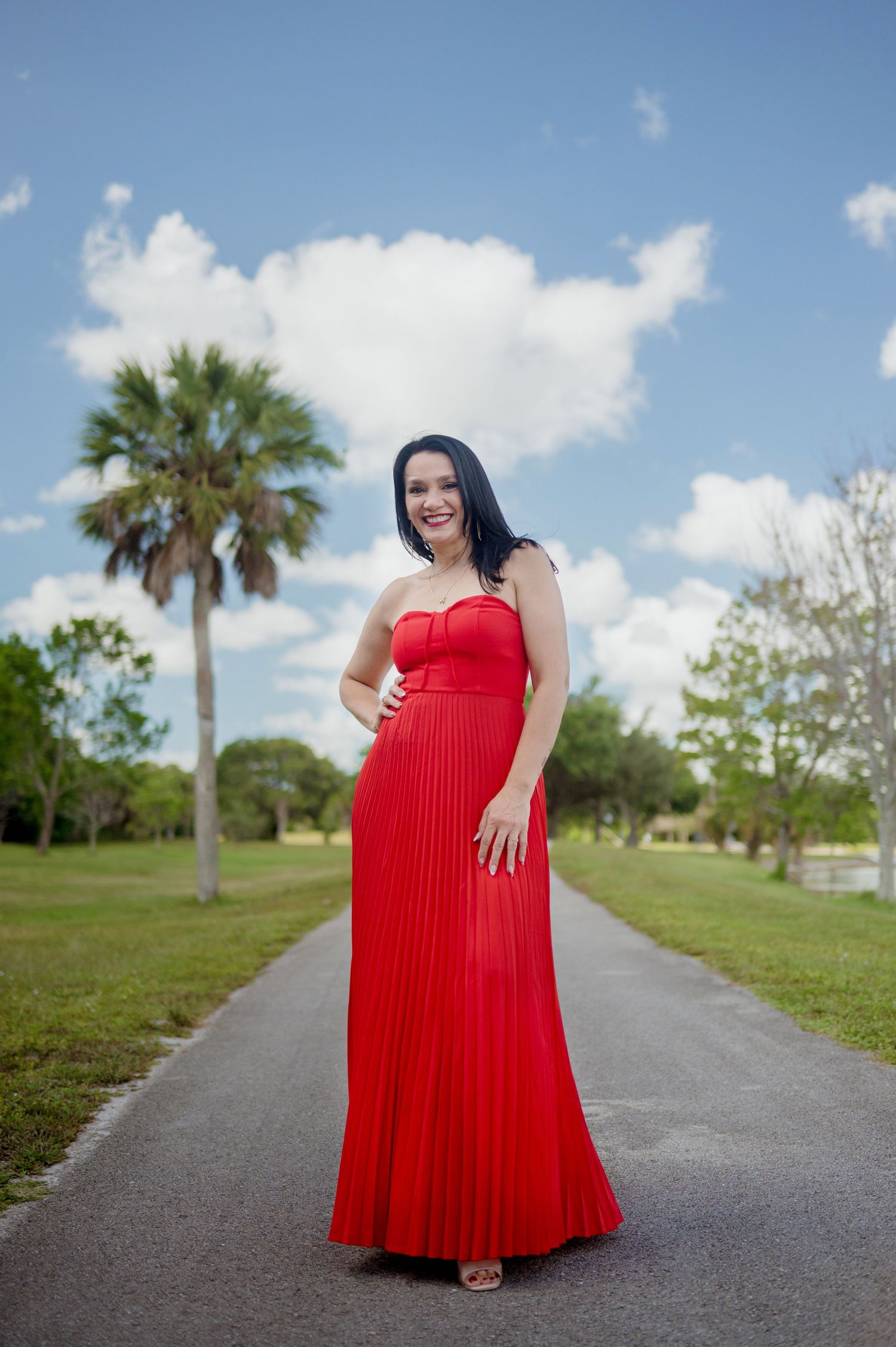 A strapless red pleated maxi dress photographed outdoors on a path with palm trees and clouds in the background.