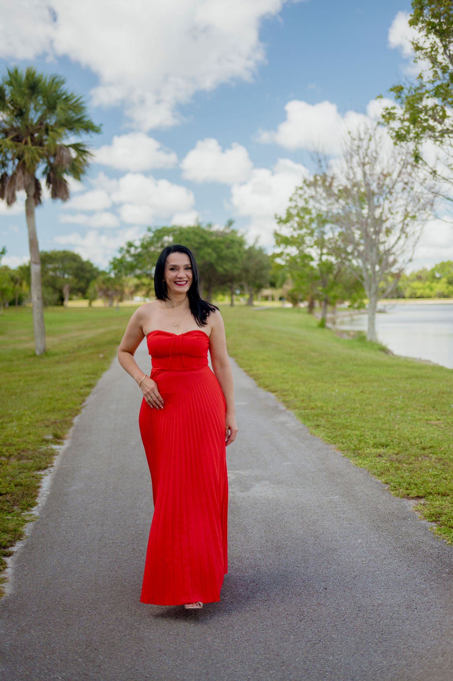 A person in a vibrant red strapless maxi dress poses on a palm tree-lined path beside a lake on a sunny day.