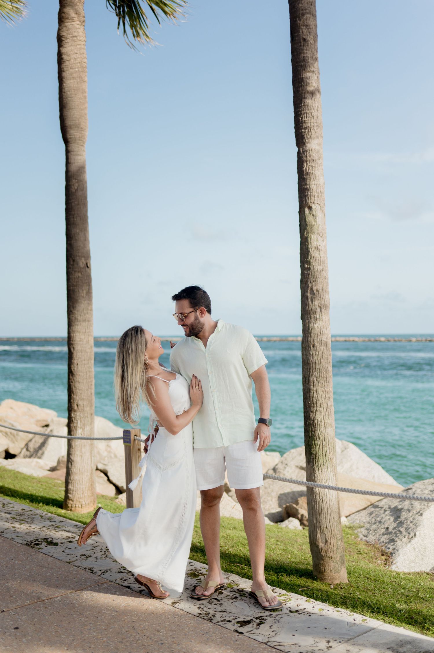A couple in white outfits stands between palm trees on a tropical beach overlooking turquoise waters.