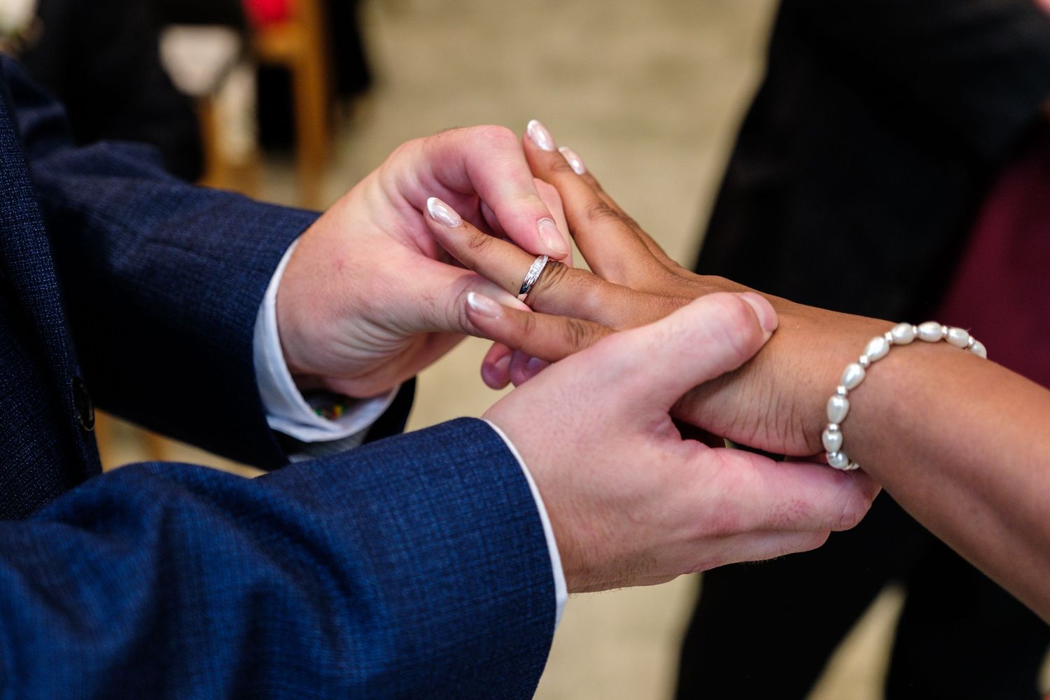 Close-up of hands exchanging wedding rings during a marriage ceremony with the groom wearing a navy blue suit.