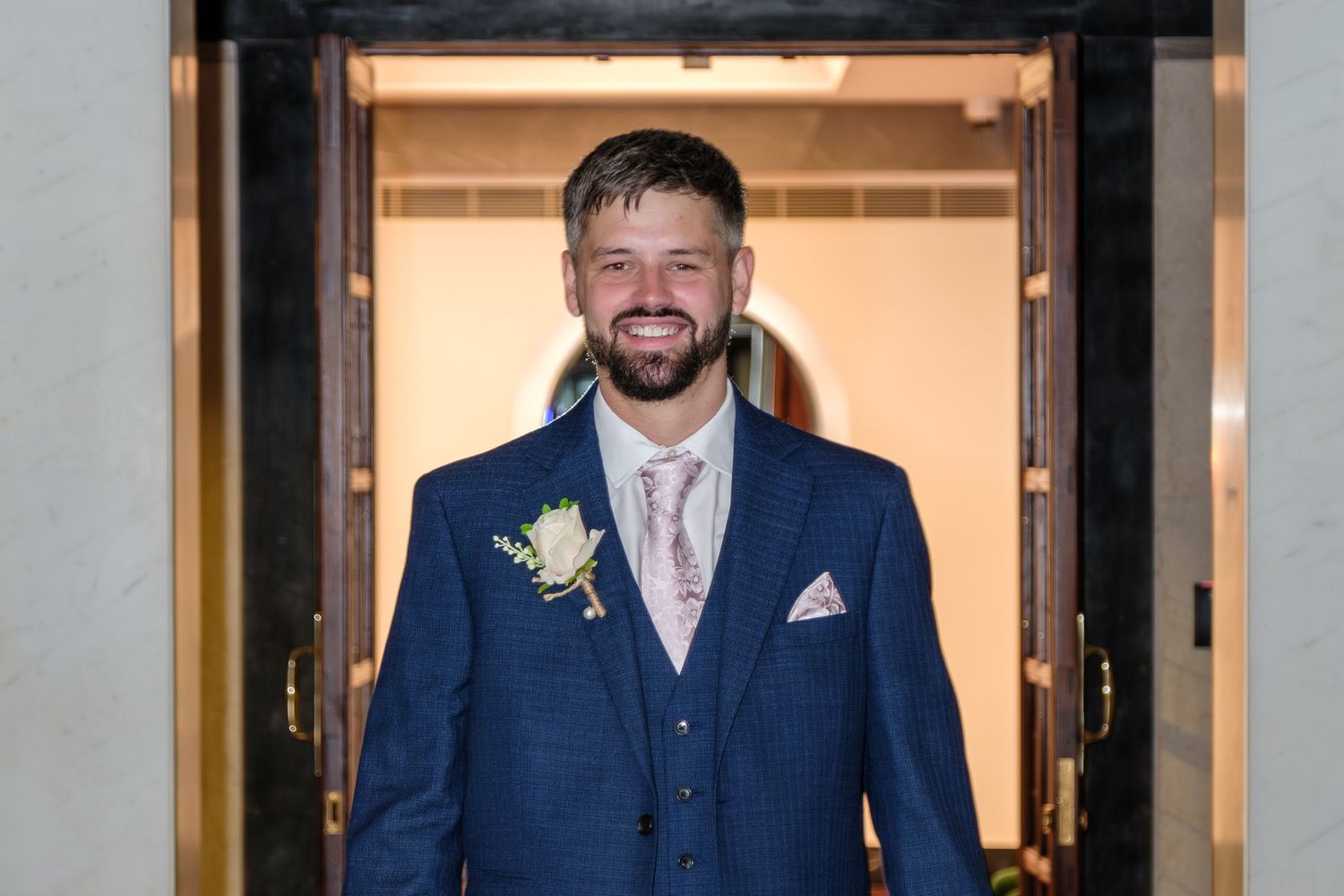 A person in a dark blue suit stands in front of illuminated glass doors at night.