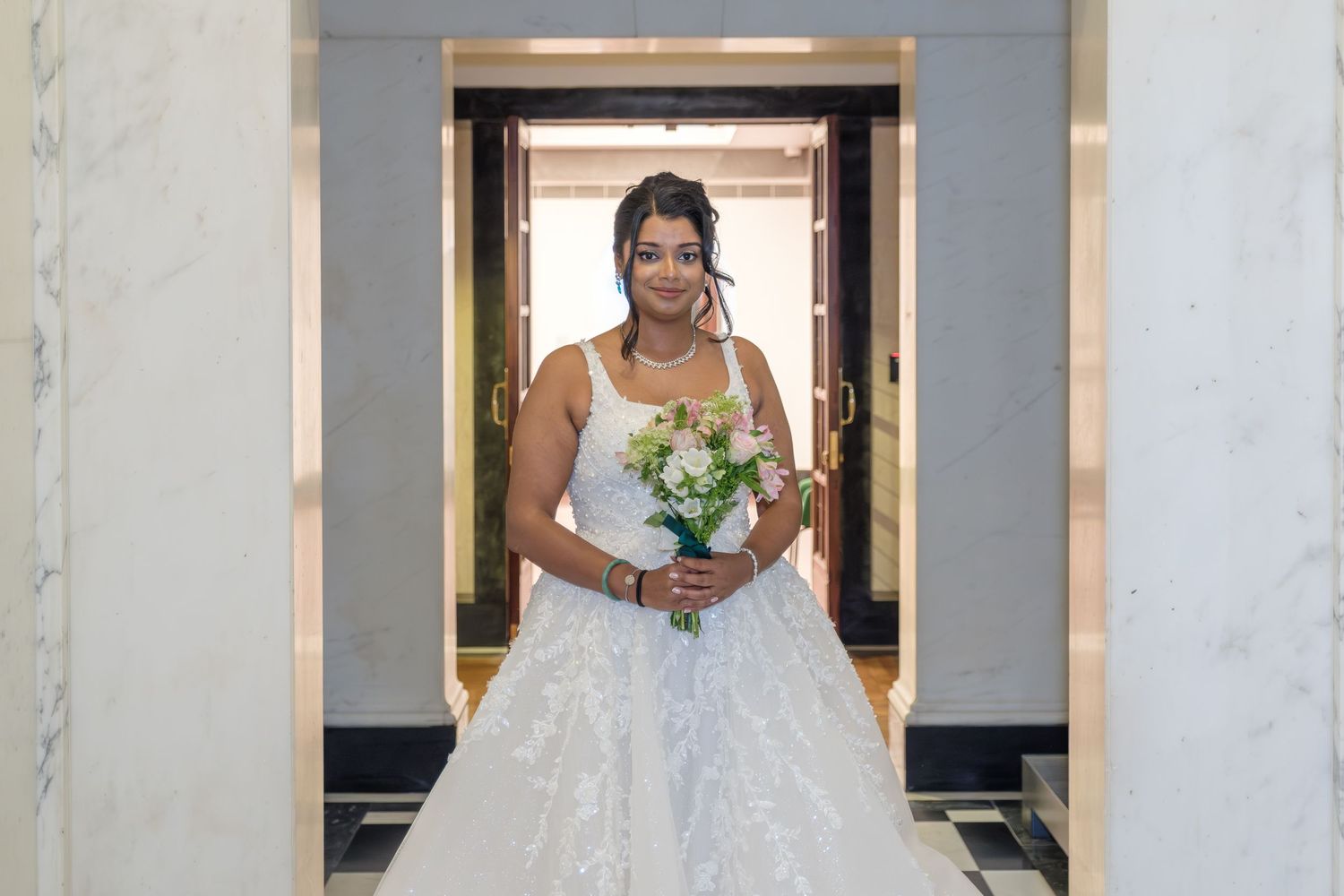 Elegant bride in white wedding dress posing against marble wall in hotel hallway with classic decor.